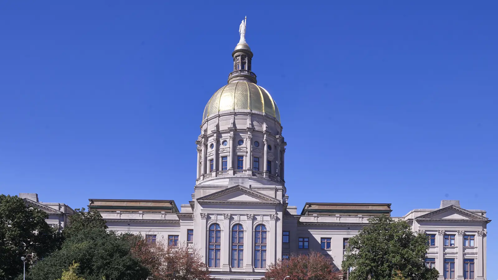 The Georgia State Capitol in Atlanta, its gold dome topped by a white statue above a pale stone facade with tall arched windows, framed by trees under a clear blue sky.